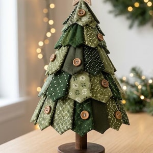 Peut inclure: Un sapin de Noël décoratif en tissu composé de segments de tissu vert à motifs dans différentes nuances, agrémenté de boutons en bois marron. L'arbre est surmonté d'une étoile en bois clair et repose sur un socle rond en bois.