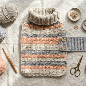 May include: A crocheted hot water bottle cover with a ribbed turtleneck, featuring horizontal stripes in cream, peach, and gray. Includes three balls of yarn, a wooden crochet hook, a sewing needle, scissors, a measuring tape, and wooden buttons.