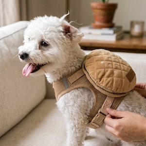 Può includere: Un piccolo cane bianco indossa una pettorina in rete color marrone chiaro con uno zainetto rotondo e trapuntato dello stesso colore. La pettorina è dotata di cinghie regolabili e di una piccola toppa rettangolare sul lato dello zaino. Il cane ha la lingua di fuori.