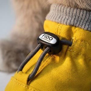 Könnte beinhalten: Ein leuchtend gelber Haustierschuh mit grauem Rippbündchen und einem schwarzen Kordelstopper, auf dem der weiße Schriftzug FIDO ACCESSORIES steht. Der Schuh wird mit einer grauen elastischen Kordel befestigt, die durch den schwarzen Stopper verläuft.