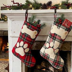 May include: Two Christmas stockings made of plaid fabric, one primarily red and one primarily white, each featuring a beige felt bone appliqué at the top and a beige felt paw print appliqué below, filled with artificial pine branches and berries.