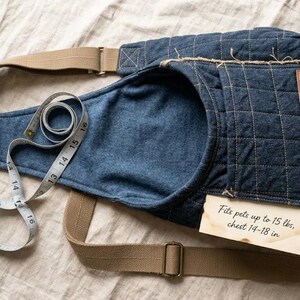 May include: A quilted blue denim pet carrier sling with tan adjustable straps and a rectangular tan leather label. A white measuring tape is coiled on the sling, and a paper tag reads: Fits pets up to 15 lbs, chest 14-18 in.