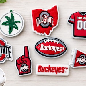 May include: A collection of Ohio State University themed shoe charms featuring a green buckeye leaf, the state of Ohio, a red jersey with 00, the Ohio State logo, a red foam finger, a football with Buckeyes text, a pennant, and a Buckeyes logo.