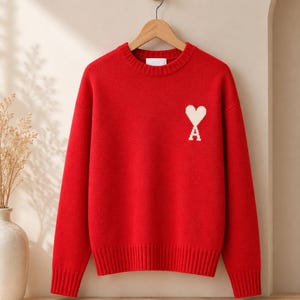 Puede incluir: Un suéter rojo vibrante de manga larga con cuello redondo, que presenta cuello, puños y dobladillo acanalados. En la parte superior izquierda del pecho, hay un corazón blanco bordado con la letra A centrada en la base, colgado en una percha de madera.