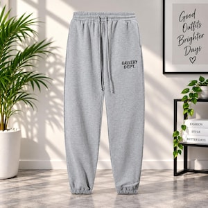 Puede incluir: Pantalones de chándal de color gris claro con cintura elástica, cordón ajustable y puños en los tobillos. El texto GALLERY DEPT. está impreso en negro en la pierna derecha. Estos pantalones tienen un corte holgado y un color gris sólido.