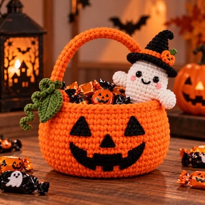 Puede incluir: Una cesta de ganchillo de color naranja brillante con forma de calabaza de Halloween y una cara negra cosida. Un pequeño fantasma blanco de ganchillo con un sombrero de bruja negro se sienta en la cesta, llena de dulces y golosinas envueltos.