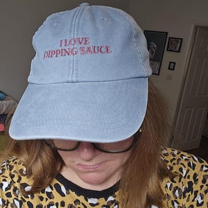 Peut inclure: Une casquette de baseball bleu clair avec une visière incurvée et une construction à six panneaux. L'avant de la casquette présente le texte brodé I LOVE DIPPING SAUCE en fil rouge. Le tissu a un aspect légèrement délavé et usé.