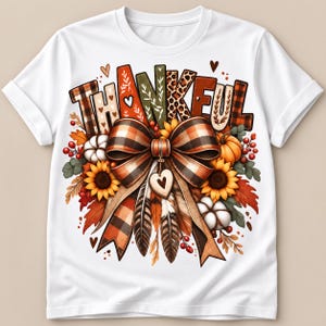 May include: A white t-shirt featuring a graphic print of the word THANKFUL in varied patterns above a large plaid bow. The design includes sunflowers, pumpkins, cotton bolls, autumn leaves, red berries, and feathers with a small heart charm.