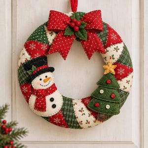 May include: A quilted fabric wreath featuring a patchwork pattern of red, green, and cream sections with snowflake and holly prints. It is decorated with a red polka-dot bow, a felt snowman wearing a black top hat, and a green felt Christmas tree.
