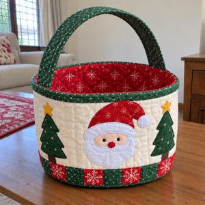 May include: A quilted fabric basket featuring a central Santa Claus face applique, flanked by two green Christmas trees with yellow stars. The basket has a cream-colored exterior, a red interior with white snowflakes, and a green handle.