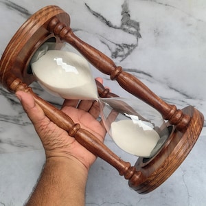 May include: An hourglass featuring a dark brown wooden frame with turned pillars and circular top and bottom bases. The clear glass bulb contains fine white sand, with the majority of the sand currently resting in the upper chamber of the device.