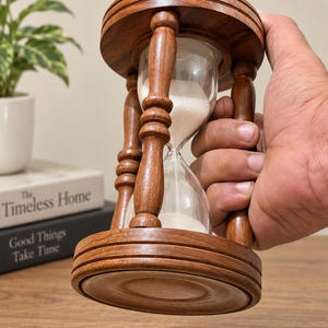 May include: An hourglass with a dark brown wooden frame featuring turned pillars and circular top and bottom bases. The clear glass bulb contains fine white sand, with a portion of the sand currently resting in the bottom chamber of the device.