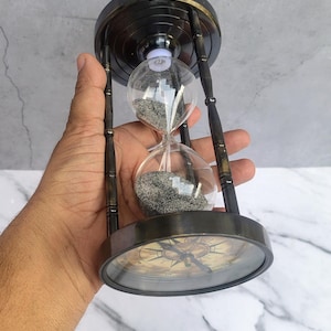 Peut inclure: Un sablier avec un cadre métallique sombre et des ampoules en verre transparent contenant du sable gris. La base du sablier comporte une boussole intégrée avec un cadran circulaire, des marquages noirs et une aiguille indiquant les points cardinaux.