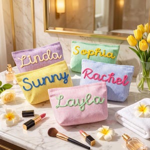 Könnte beinhalten: Vier gestreifte Kosmetiktaschen in Lila, Gelb, Grün und Pink, jeweils mit einem Namen in erhabener Schreibschrift: Linda, Sunny, Sophia, Rachel und Layla. Die Taschen sind mit Make-up-Pinseln, Lippenstiften und Blumen arrangiert.