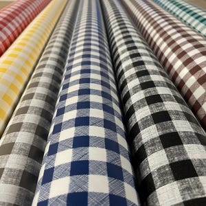 Può includere: Diversi rotoli di tessuto disposti fianco a fianco, ognuno caratterizzato da un classico motivo a quadretti vichy. I colori includono rosso, giallo, grigio, blu navy, nero, marrone e verde acqua su uno sfondo bianco con trama tessuta visibile.