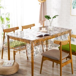 Puede incluir: Una mesa de comedor rectangular cubierta con un mantel de plástico transparente que presenta un patrón floral de líneas negras. La mesa es de madera clara y está rodeada por cuatro sillas de madera con cojines de tela verde y gris.