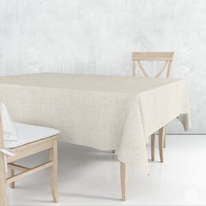 Könnte beinhalten: Eine rechteckige Tischdecke in neutralem Beige mit einer sichtbaren Webstruktur. Die Tischdecke liegt auf einem Tisch, der von zwei hellen Holzstühlen mit weißen Sitzkissen in einem hellen Raum begleitet wird.