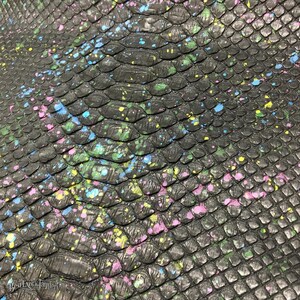 May include: A black snakeskin leather sheet featuring a textured scale pattern with scattered paint splatters in vibrant shades of blue, green, pink, and yellow. The text BestExoticTannery is printed in the bottom left corner.
