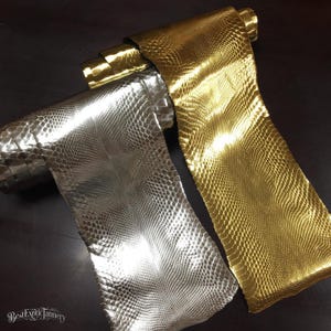 Puede incluir: Dos rollos de cuero metálico con un patrón texturizado de piel de serpiente. Un rollo tiene un acabado en tono plateado reflectante, mientras que el otro tiene un acabado en tono dorado reflectante. El texto BestExoticTannery aparece en la esquina.