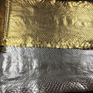 May include: Two pieces of textured animal skin leather are displayed. The top piece is a metallic gold color with a distinct scale pattern. The bottom piece is a metallic silver color with a similar scale pattern. The text Best Exotic Tannery is visible.