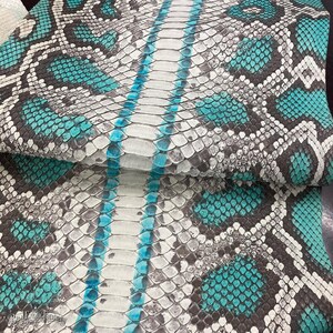 May include: A sheet of snakeskin leather featuring a repeating pattern of turquoise, white, and dark gray scales. A central vertical band of larger, elongated white scales runs down the middle, accented by bright turquoise edges and texture.