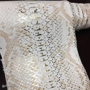 May include: A roll of white and beige snakeskin leather featuring a metallic gold finish on the scales. The material displays a repeating diamond pattern with a textured surface. The text Best Exotic Tannery is printed in the bottom corner.