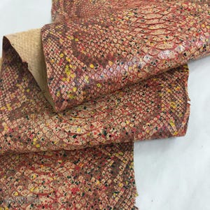 May include: A piece of textured snakeskin leather featuring a multi-colored pattern with shades of red, brown, and tan, accented by small, irregular splatters of bright yellow, green, and black paint across the scale-like surface texture.