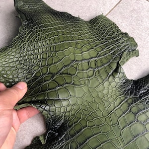 Puede incluir: Una pieza de cuero verde oliva con un patrón texturizado y en relieve que se asemeja a escamas de reptil. La superficie presenta un degradado de sombreado negro oscuro en las grietas del patrón. El texto BestExoticTannery es visible.