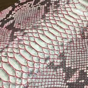 May include: A piece of snakeskin leather featuring a repeating scale pattern in shades of white, gray, and black, with vibrant red accents outlining the edges of the individual scales. The texture is uniform and detailed across the surface.