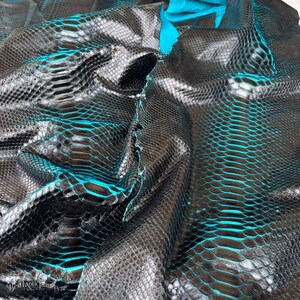 May include: A piece of dark brown snakeskin leather featuring a distinct scale pattern with vibrant turquoise blue accents visible between the scales. The material is folded, creating deep shadows and highlights across the textured surface.