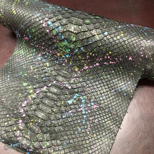 May include: A rolled piece of dark gray textured leather featuring a repeating scale pattern. The surface is decorated with scattered paint splatters in vibrant shades of neon pink, bright yellow, and light blue across the entire material.