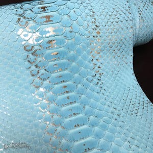 May include: A light blue snakeskin leather sheet featuring a repeating scale pattern with metallic silver accents throughout the surface. The bottom left corner displays the text BestExoticTannery in a small, stylized font.