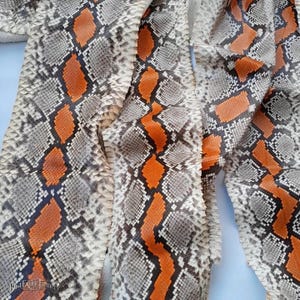 May include: A piece of snakeskin leather featuring a repeating diamond pattern in vibrant orange, grey, and white scales. The texture is detailed and organic, with the text Best Exotic Tannery visible in the bottom left corner of the material.