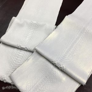 以下が含まれることがあります： 表面全体に独特の隆起した鱗模様が施された、複数の白いヘビ革。素材はマットな質感で、ほのかな光沢があります。画像左下隅にはBestExoticTanneryというテキストが印刷されています。