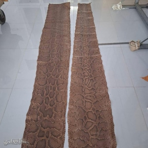 May include: Two long, flat strips of brown snakeskin leather laid out vertically. The material features a natural, repeating diamond-shaped scale pattern in shades of tan and dark brown. The text BestExoticTannery is visible in the bottom left corner.