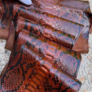 May include: A stack of multiple sheets of glossy, reddish-brown snakeskin leather featuring a distinct, overlapping scale pattern with dark brown and black accents. The text BestExoticTannery is visible in the bottom left corner of the image.