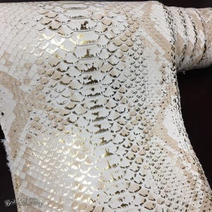 Puede incluir: Un rollo de piel de serpiente en color blanco y oro metálico con un patrón de escamas repetitivo. El material presenta una superficie texturizada con un acabado reflectante en las escamas, creando un contraste entre los tonos blancos y dorados.