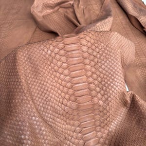 May include: A large, folded piece of light brown snakeskin leather featuring a distinct, repeating scale pattern across the entire surface. The texture is uniform with a matte finish. The text BestExoticTannery is visible in the corner.