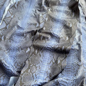 May include: A large piece of blue and grey snakeskin leather featuring a natural scale pattern and a textured surface. The material displays a repeating diamond-like design with a gradient of cool tones. The text BestExoticTannery is visible.