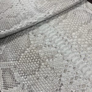 Könnte beinhalten: Ein gefaltetes Stück silberfarbener Stoff mit einer strukturierten Schlangenhaut-Optik. Das Material weist einen metallischen Glanz mit variierenden Schuppen auf der Oberfläche auf. Der Text BestLeatherTannery steht unten links im Bild.