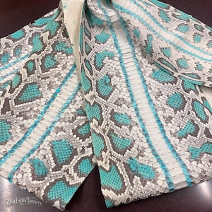 May include: A piece of snakeskin leather featuring a repeating scale pattern in shades of white, gray, and bright turquoise. The material displays a natural texture with a central row of larger scales and the text Best Exotic Tannery.