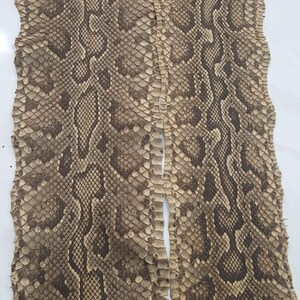 May include: A long, flat piece of snakeskin leather featuring a natural, repeating diamond-shaped scale pattern in shades of tan, beige, and dark brown. The surface has a textured, organic appearance with the text BestExoticTannery visible at the bottom.