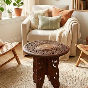 Può includere: Un tavolino rotondo in legno marrone scuro con intricati intagli floreali sul piano e una base pieghevole con dettagli intagliati. Il tavolo presenta un inserto centrale circolare e poggia su quattro gambe intagliate che sostengono il piano.