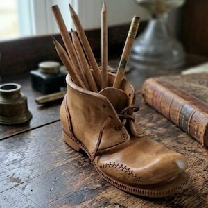 Puede incluir: Un portalápices con forma de zapato de cuero color canela con costuras visibles en el lateral y cordones en la parte superior. El zapato contiene varios lápices de madera con puntas de grafito, incluido uno con virola metálica.