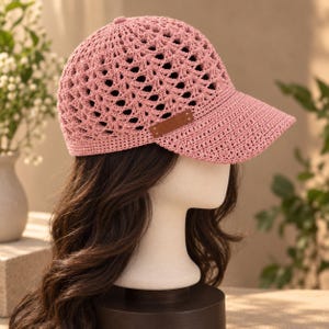 May include: A dusty pink baseball cap crafted from a crocheted yarn material featuring an openwork fan pattern across the crown. The cap includes a solid brim and a small rectangular brown faux leather patch detail positioned on the side panel.