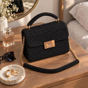 May include: A black crocheted handbag featuring a structured rectangular shape, a top handle, and a gold-tone metal chain shoulder strap. The bag is secured with a rectangular gold-tone metal push-lock clasp centered on the front flap.