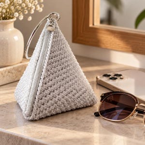 Pode incluir: Uma bolsa triangular de crochê em cinza claro com um padrão de pontos texturizado, um fecho de correr em metal prateado e uma alça circular em metal prateado fixada no topo. A bolsa tem uma forma geométrica estruturada.