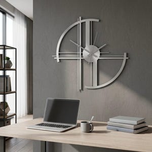 Op de afbeelding: Een zilverkleurige metalen wandklok met een open, geometrisch ontwerp met kruisende verticale en horizontale lijnen, een cirkelvormig midden en dunne wijzers, gemonteerd op een grijze muur boven een bureau met een laptop en boeken.