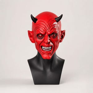 Può includere: Una maschera da diavolo rossa con corna nere, orecchie a punta ed espressione ringhiante con denti bianchi visibili. La maschera è esposta su un busto di manichino nero, mostrando rughe facciali dettagliate e occhi profondi con finitura scura.
