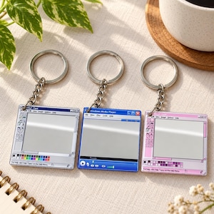 Peut inclure: Trois porte-clés carrés présentant des designs d'interface de logiciels informatiques rétro. À gauche, une fenêtre Paint grise, au centre, une fenêtre Windows Media Player bleue, et à droite, une fenêtre Paint rose. Chaque pièce a un anneau en métal argenté.